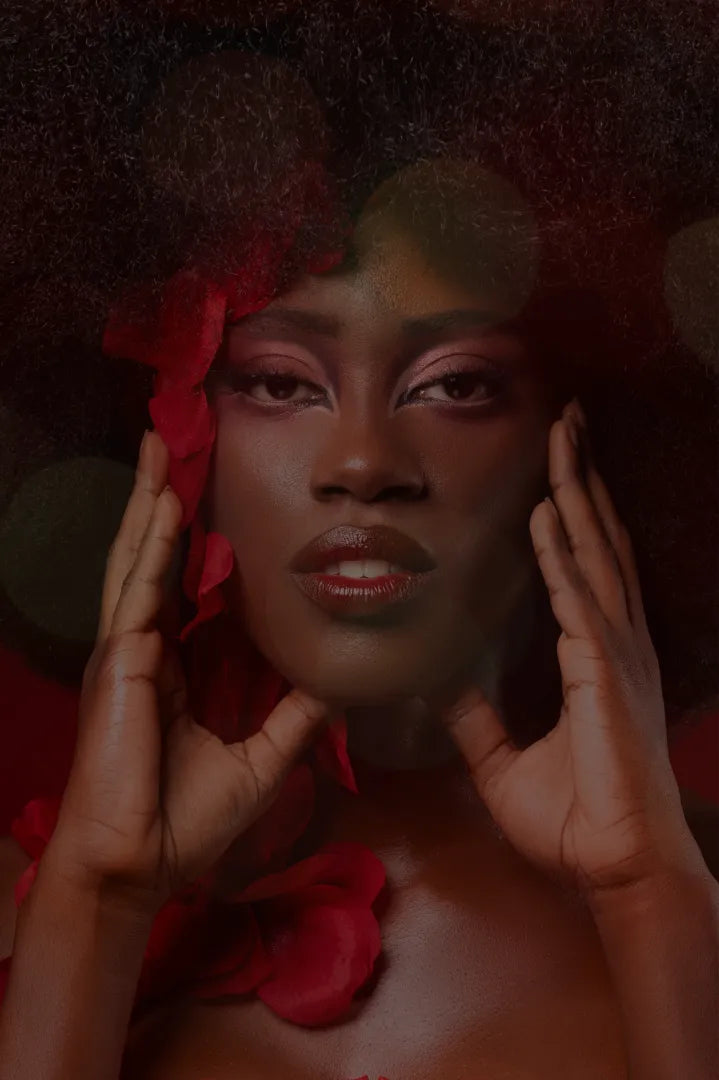 A close-up portrait of a woman with dark skin, pink eyeshadow, and red lipstick, framed by dark hair and red rose petals. Her hands are held against her cheeks, and red rose petals are scattered around her shoulders and hair against a dark, atmospheric background.